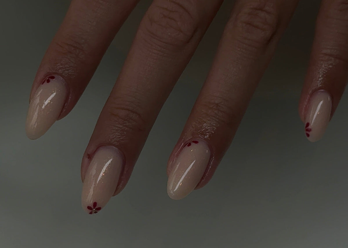 A close-up of a hand with long, almond-shaped nude nails decorated with small red floral details near the tips. A gold ring is visible on one finger, and the hand is resting on a light-colored surface.
salon apeldoorn minimale nail art clean girl nagels