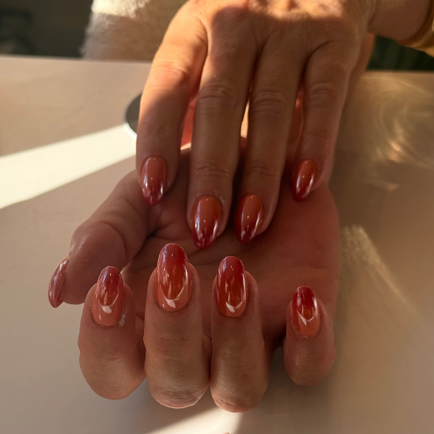 A close-up of hands with manicured nails painted in a glossy warm terracotta or rust red color, resting on a light surface with natural sunlight casting shadows. Salon Apeldoorn chrome babyboom color colorboom